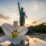 夕焼け空の下、指を天に向ける平和祈念像の後ろで、水面に映る白いユリの花が前景に置かれている。